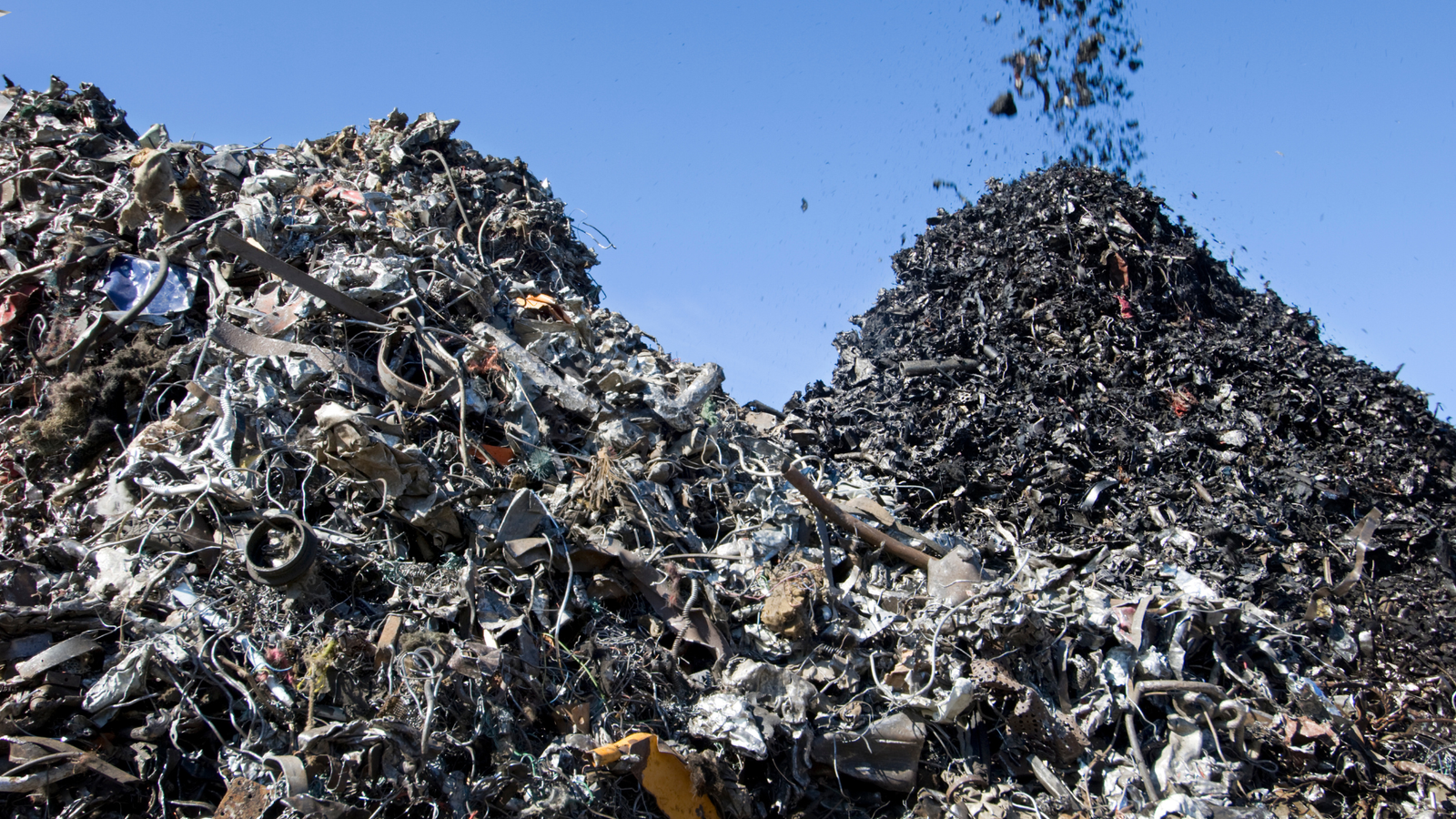 Pilhas de sucata metálica sendo processadas em um pátio industrial a céu aberto, com céu azul ao fundo. A imagem mostra diversos tipos de metais misturados, incluindo fios, chapas e peças oxidadas.