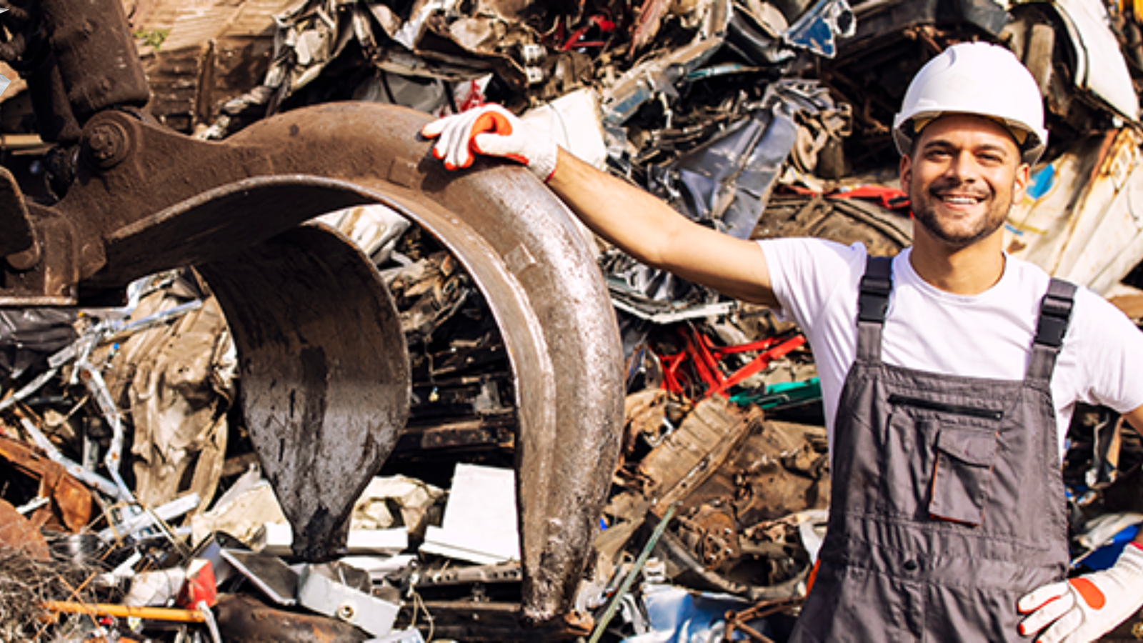 Profissional sorridente com capacete e uniforme de segurança em frente a uma pilha de sucatas metálicas, ao lado de máquina industrial, representando o setor de gerenciamento de resíduos.